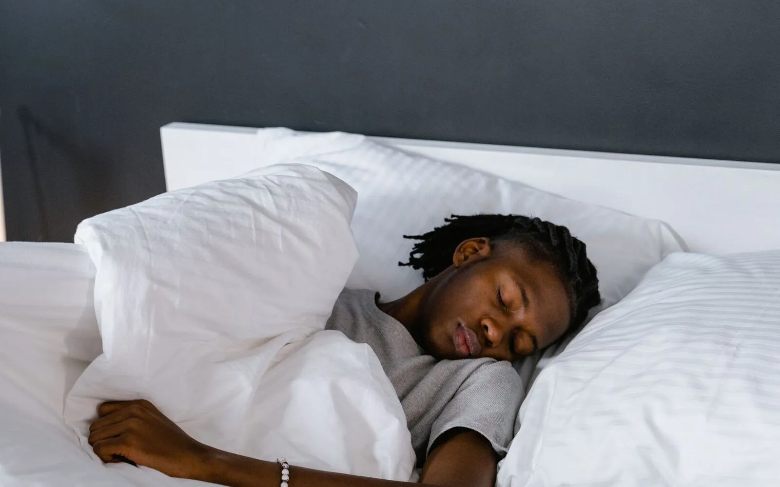A person asleep on their side in a white bed, head on the pillow, one arm resting on the duvet, against a dark grey wall.