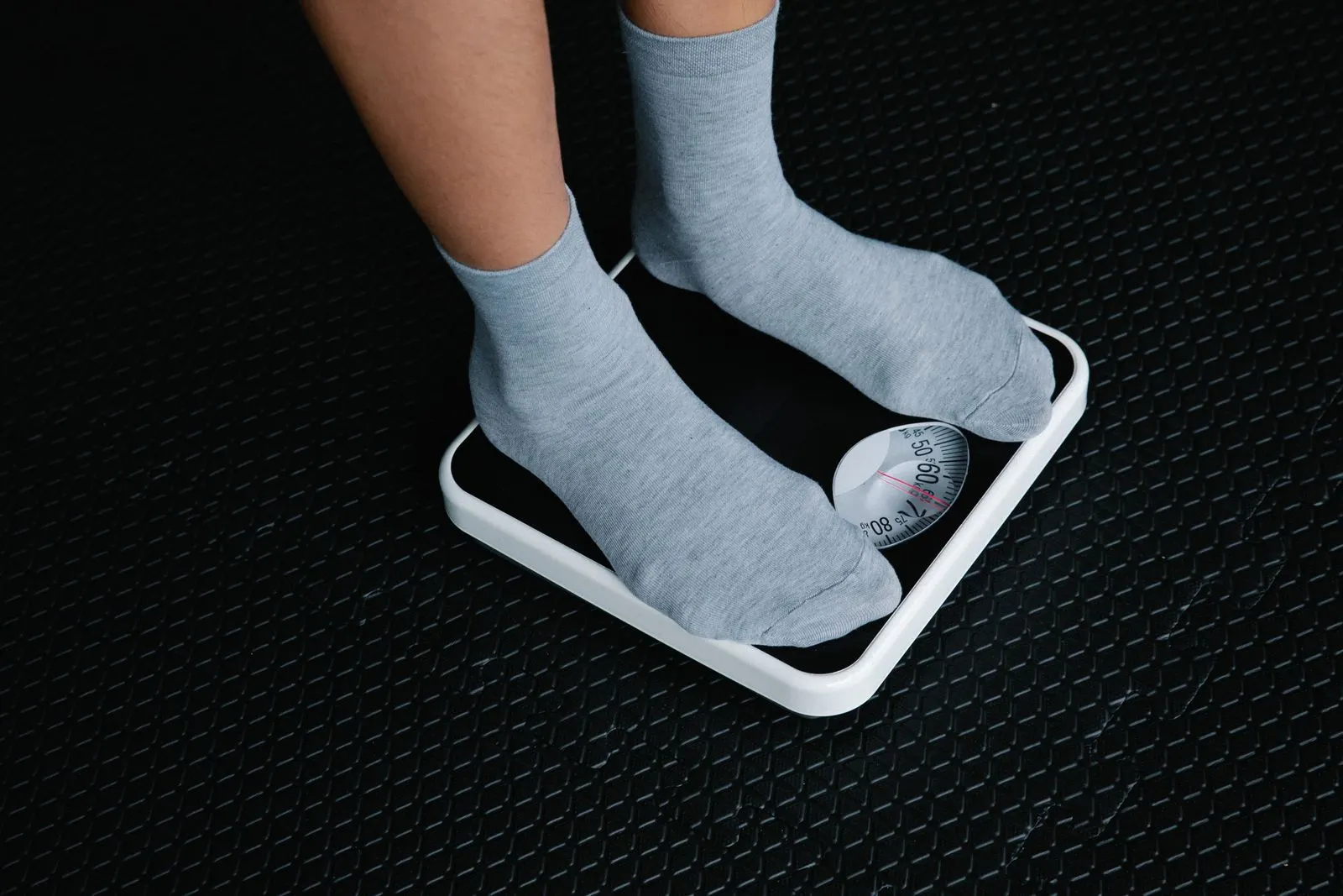Feet in grey socks standing on a white analog bathroom scale on a black rubber floor.