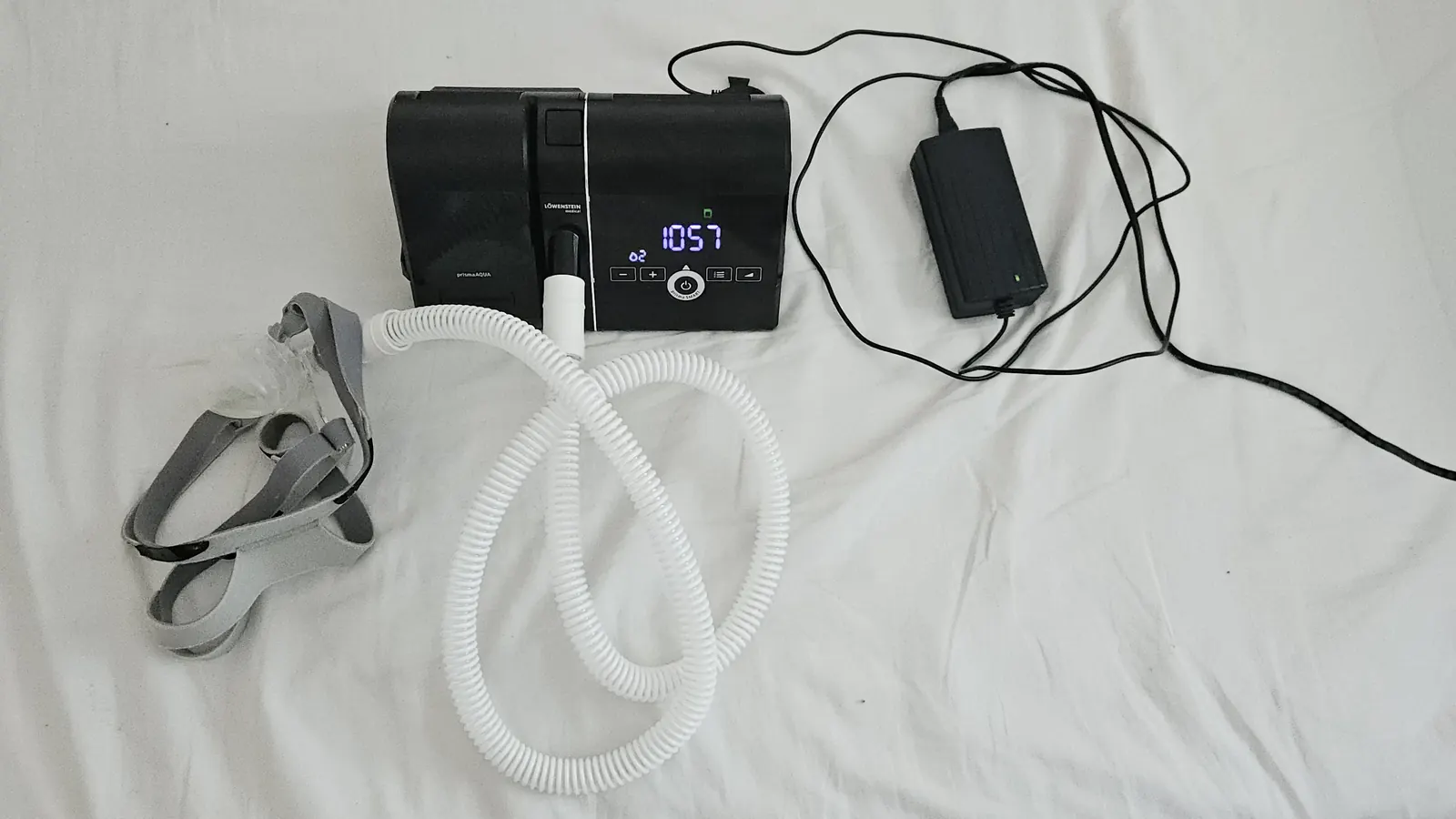 A CPAP machine with its hose, nasal mask with grey headgear, and power adapter laid out on a white bed sheet.